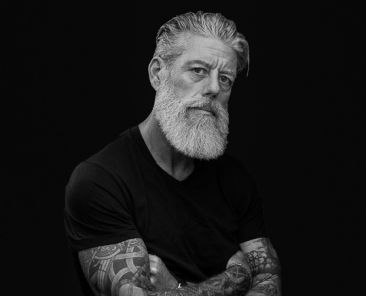 Tattooed man with his arms crossed posing in front of a black plain background.