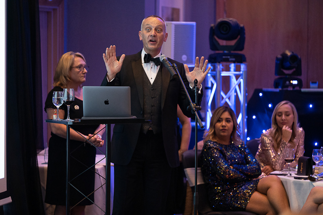 Man wearing tuxedo standing behind microphone holding his hands up looking surprised.